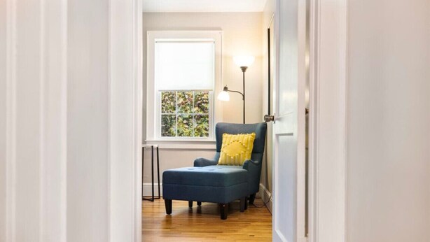 Een smalle gang met een blauwe fauteuil met een geel kussen, een vloerlamp en een raam met uitzicht op groen.