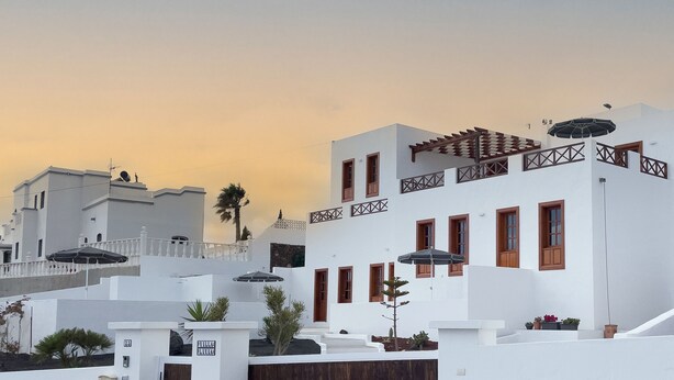 Un edificio blanco con múltiples ventanas y un balcón, rodeado de otros edificios blancos y palmeras.