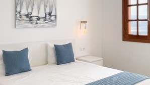 Una cama bien hecha con colcha blanca y azul, dos almohadas a juego, una lámpara fijada a la pared y una ventana con vista al mar.