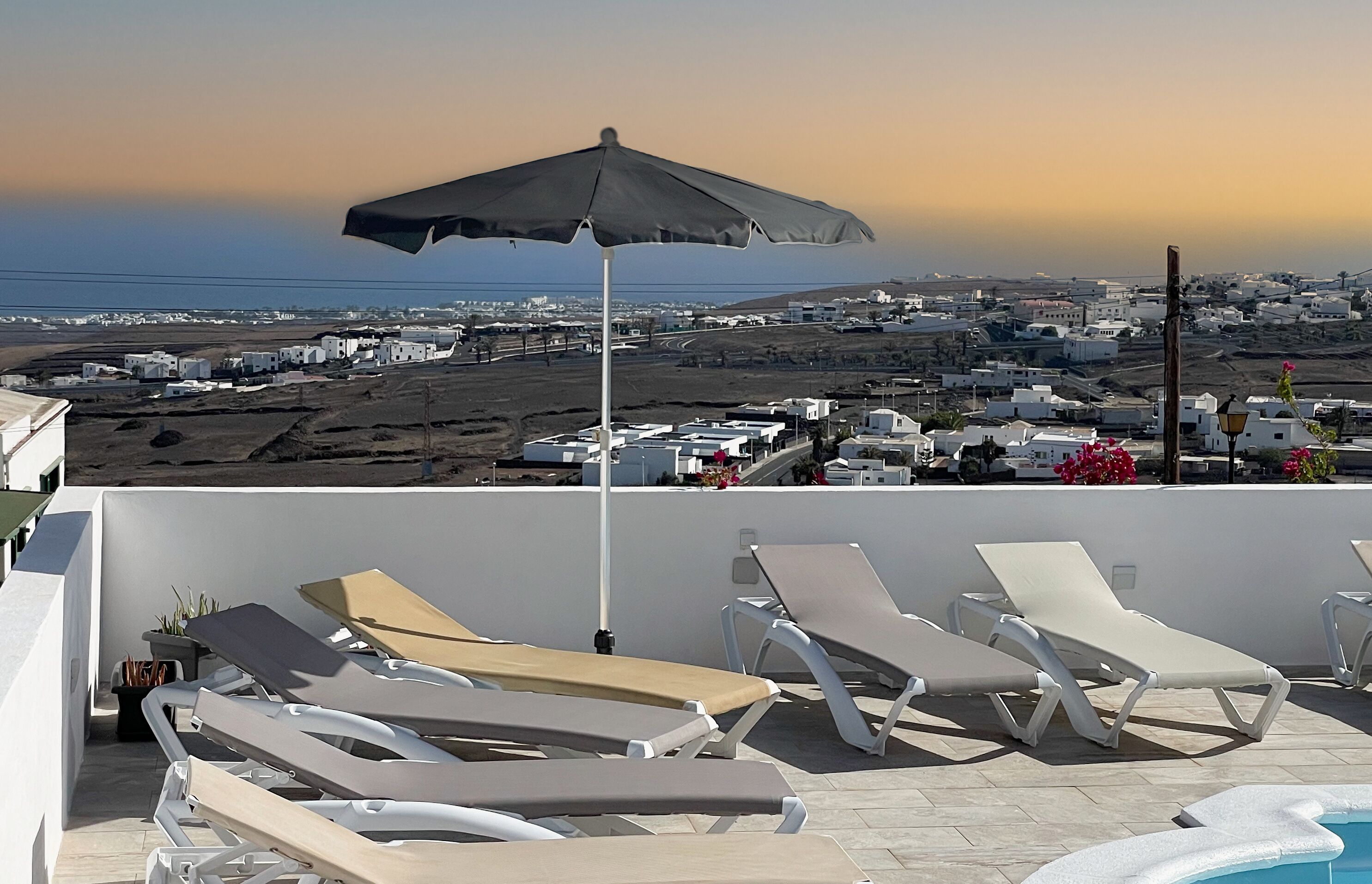 A rooftop pool area with lounge chairs and an umbrella, overlooking a coastal town.
