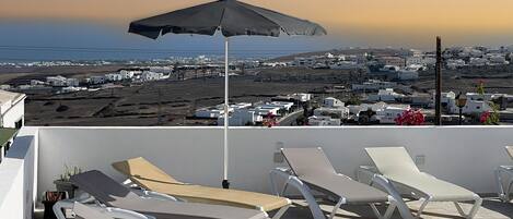 A rooftop pool area with lounge chairs and an umbrella, overlooking a coastal town.