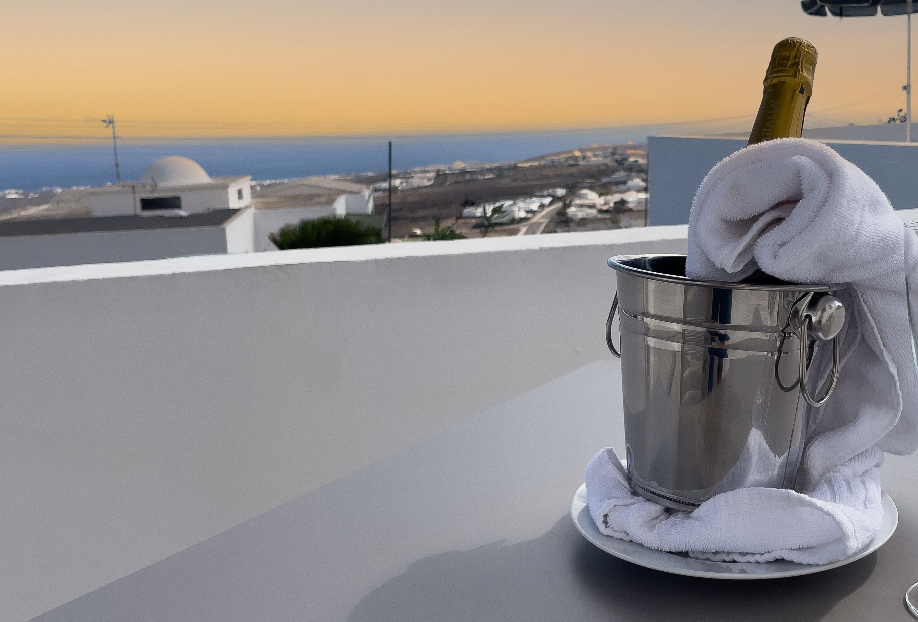 Un seau en métal, une bouteille de champagne, une serviette blanche et une assiette, le tout posé sur un balcon donnant sur un paysage urbain le long de la côte.