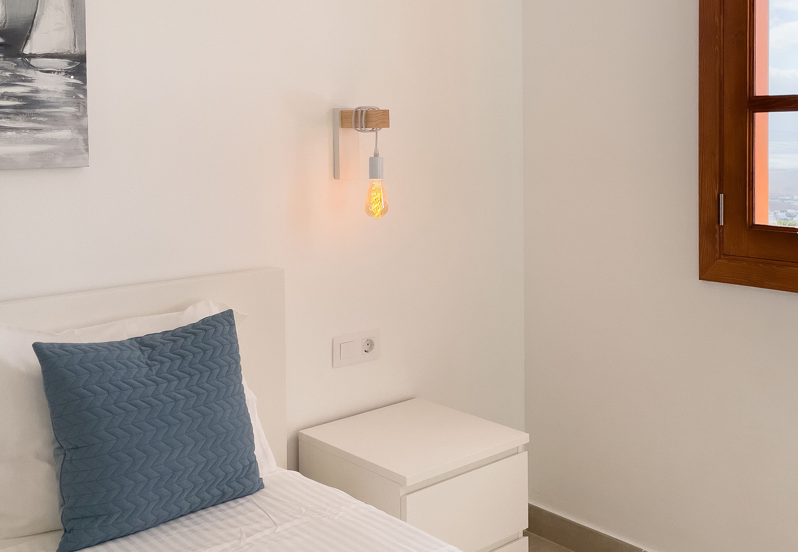 Un dormitorio con cama, mesita de noche, luz empotrada en la pared y ventana.