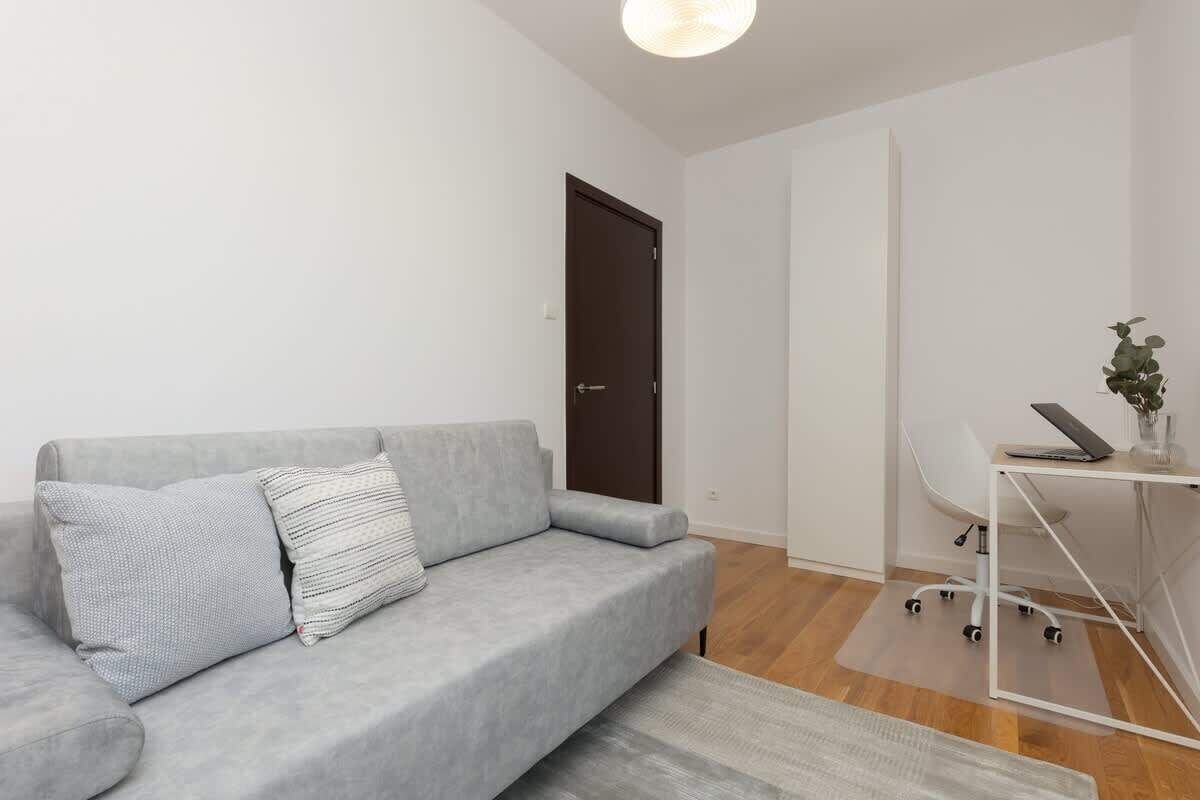 Uma sala de estar moderna com um sofá cinzento, uma pequena mesa de escritório com um laptop e uma porta.