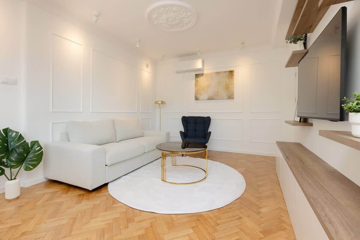 Un salon moderne avec un canapé blanc, un fauteuil foncé, une table basse ronde et un grand téléviseur à écran plat fixé au mur.
