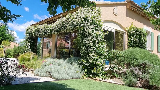 Ett hus med en trädgård, en pergola översållad med vita blommor och en klarblå himmel.
