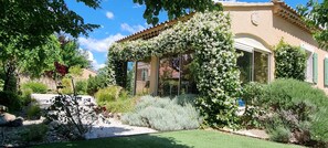 A house with a garden, a pergola covered in white flowers, and a clear blue sky.