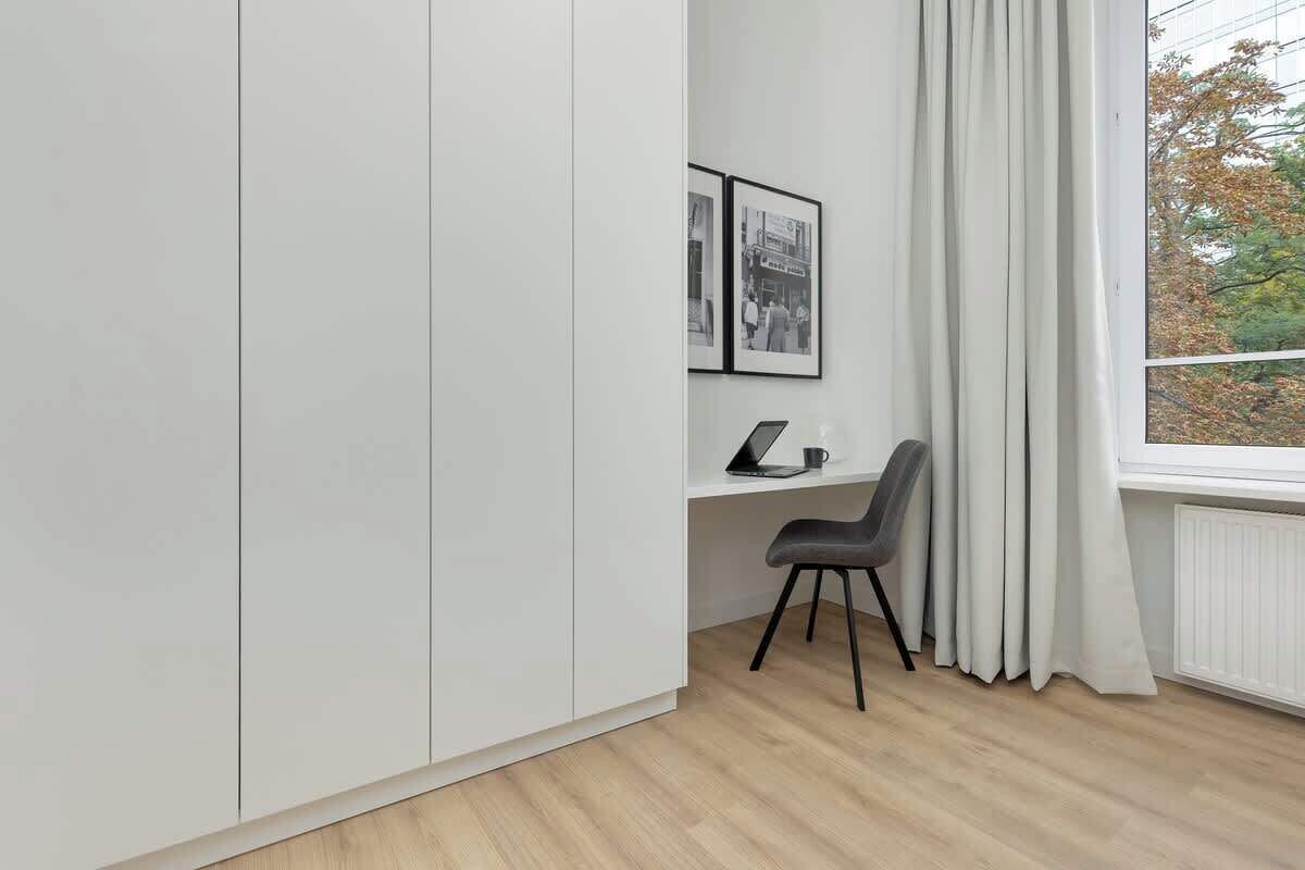 Una habitación moderna con un armario blanco, un escritorio con una computadora portátil, una silla y una ventana con cortinas.