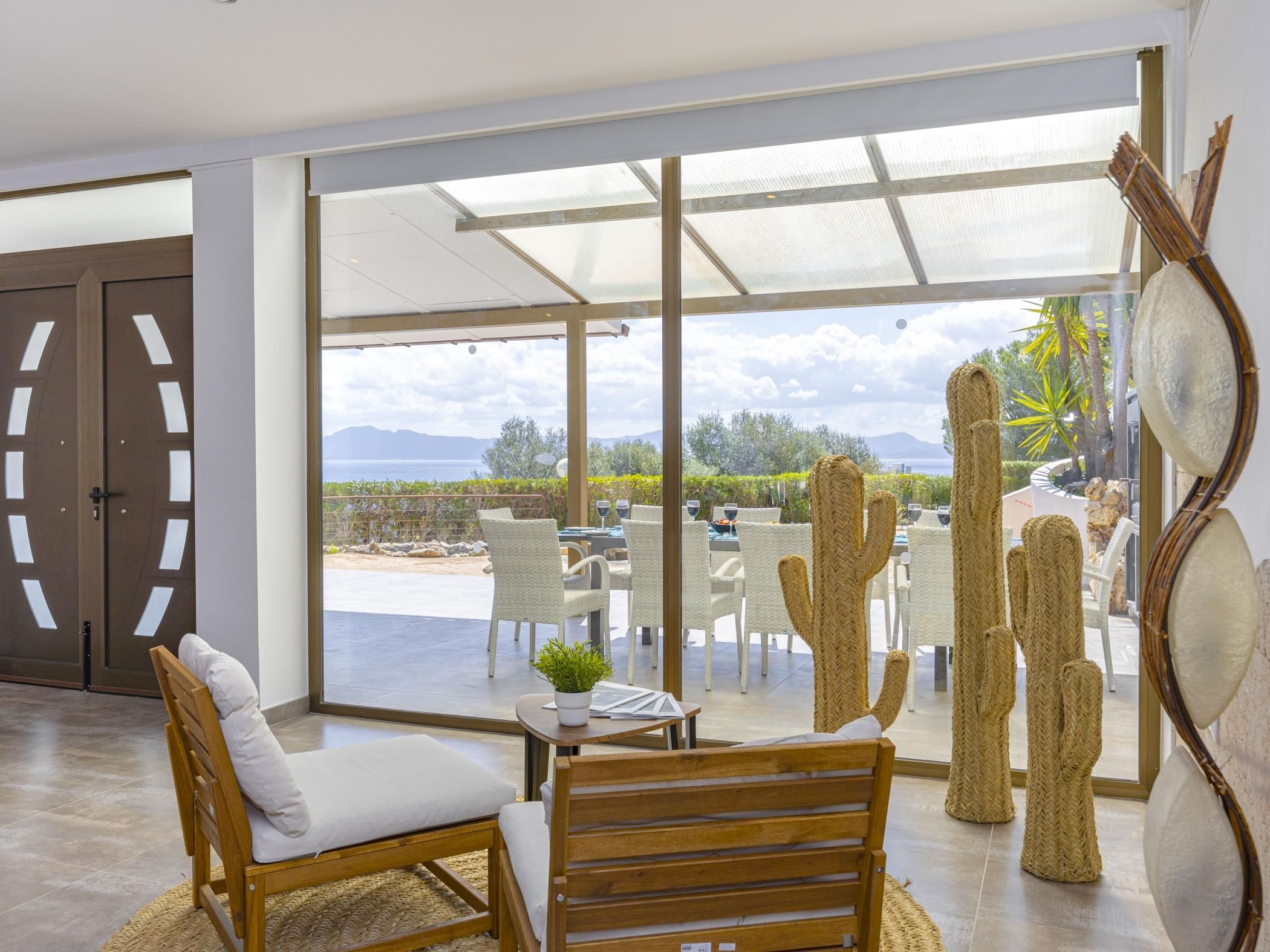 A modern room with a large sliding glass door leading to an outdoor patio with a view of the sea and mountains. There are wicker chairs and a wooden table with a plant on it.