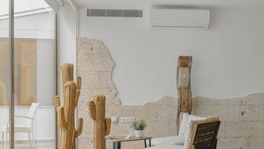 Een moderne woonkamer met een groot raam, een cactussulptuur, een rond geweven vloerkleed, een houten stoel en een wandairconditioner.