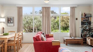 A cozy living room with a red sofa, wooden dining table, and chairs. Large windows offer a view of greenery outside.