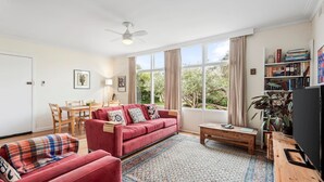 A living room with a red sofa, a wooden coffee table, a dining table with chairs, and a bookshelf.