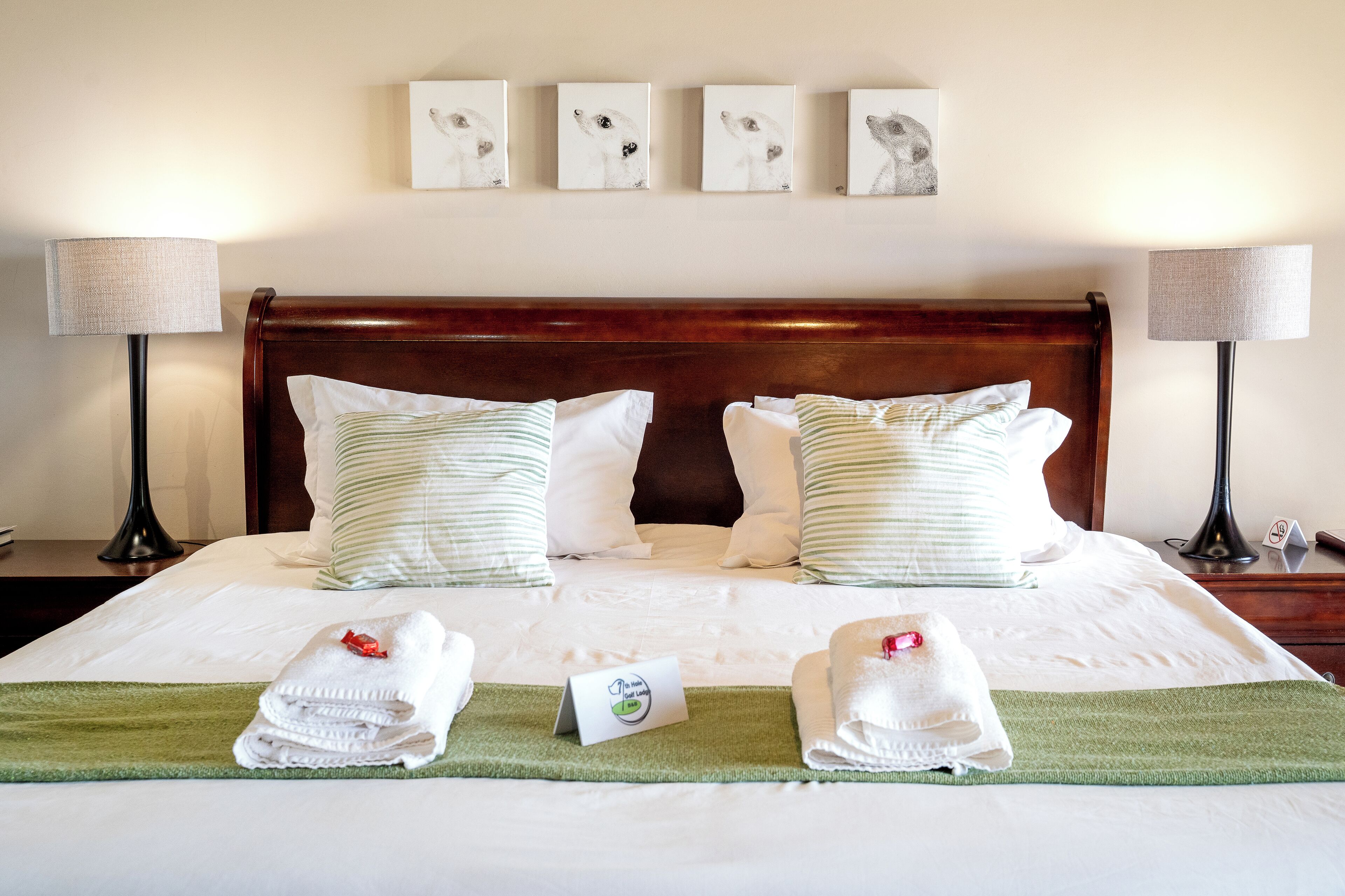 A neatly made double bed with white linens, two pillows, and a green throw blanket. There are two bedside tables with lamps and a small card on the bed.
