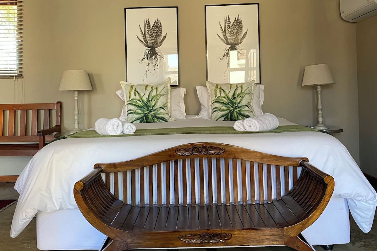 A bedroom with a bed, two framed botanical prints, a bench, and a bedside table with lamps.