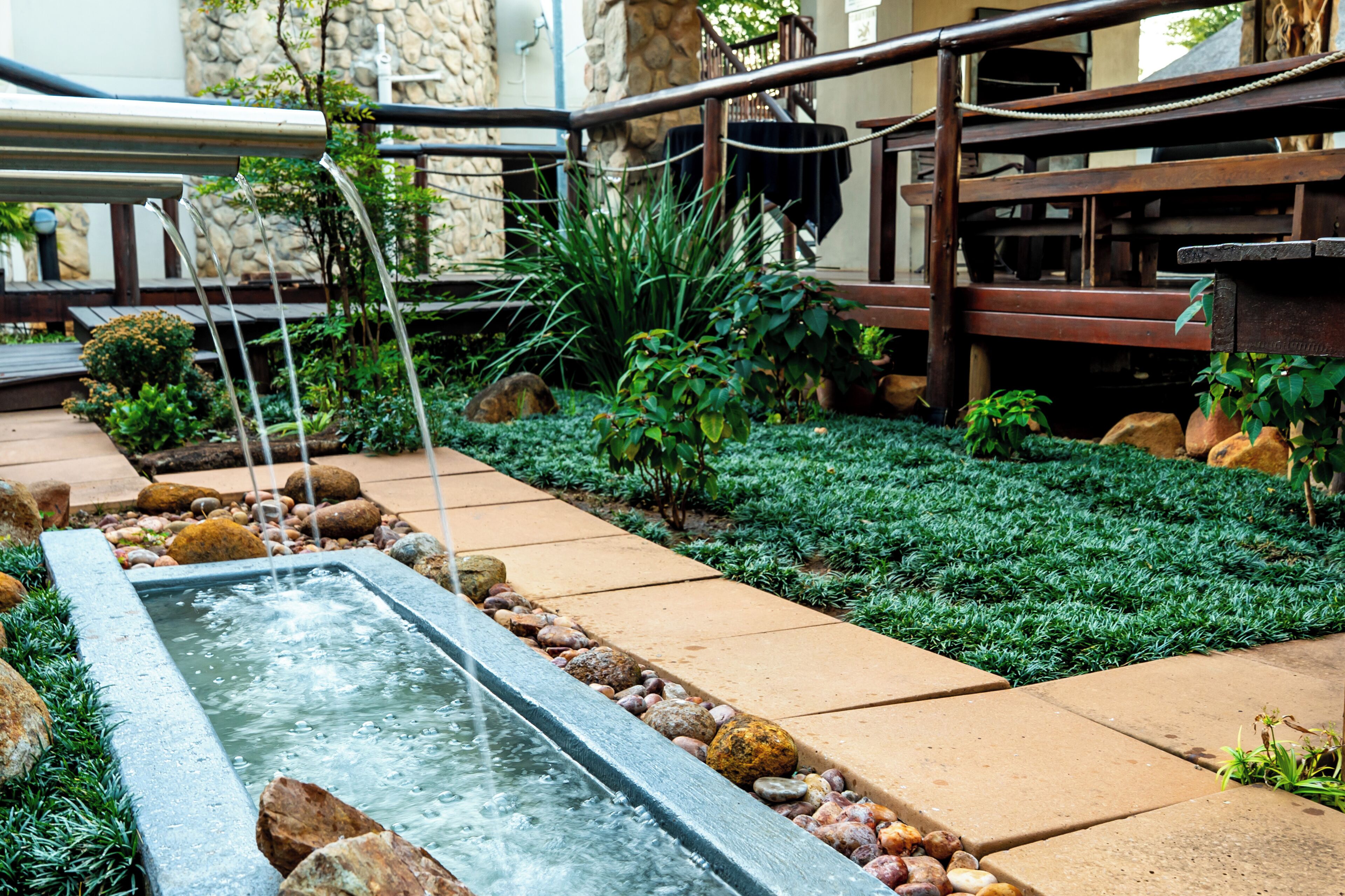 A landscaped garden with a rectangular water feature, surrounded by rocks and plants, and a wooden deck area.