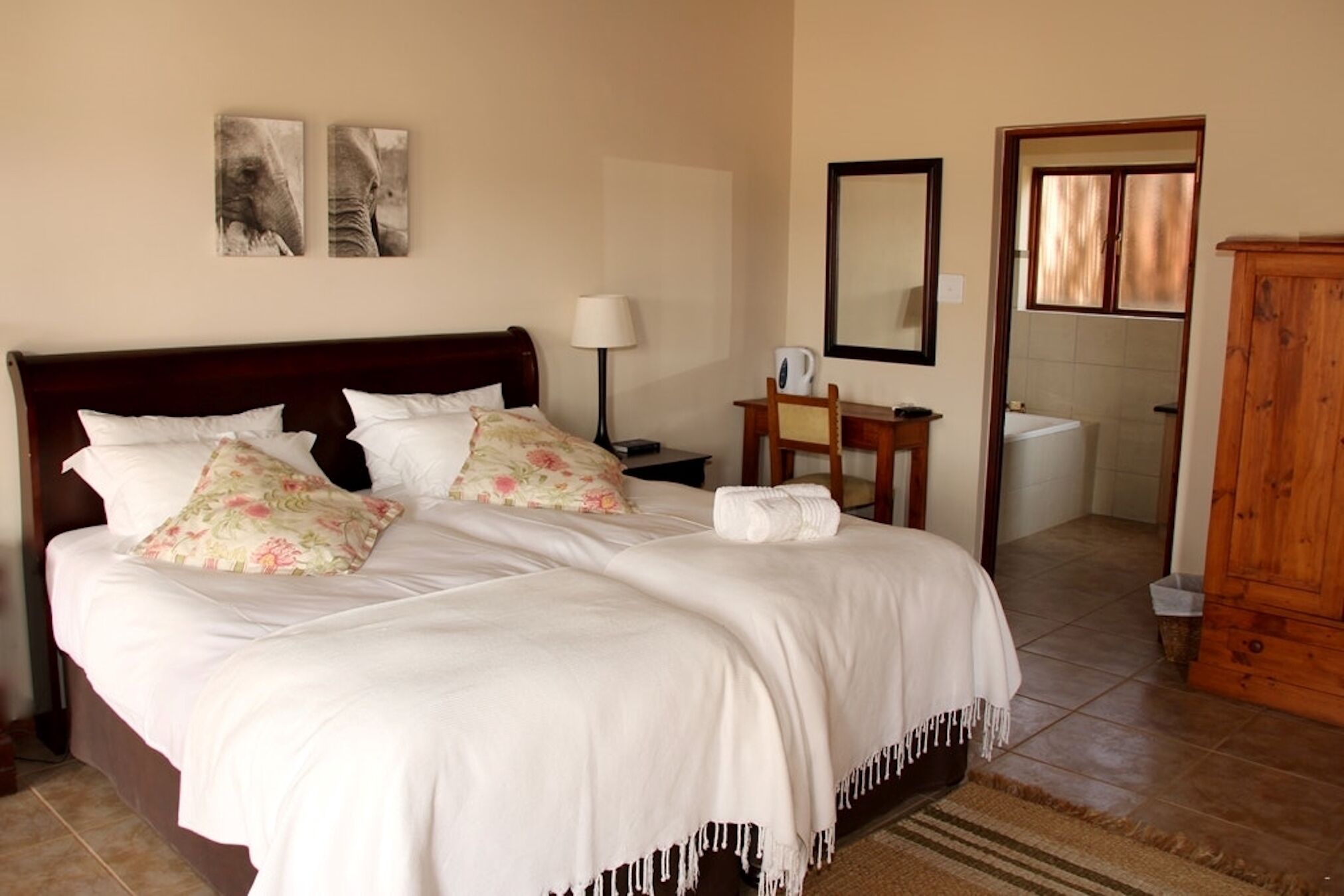 A neatly made bed with floral pillows, a bedside table with a lamp, a wooden cabinet, and a bathroom visible through an open door.