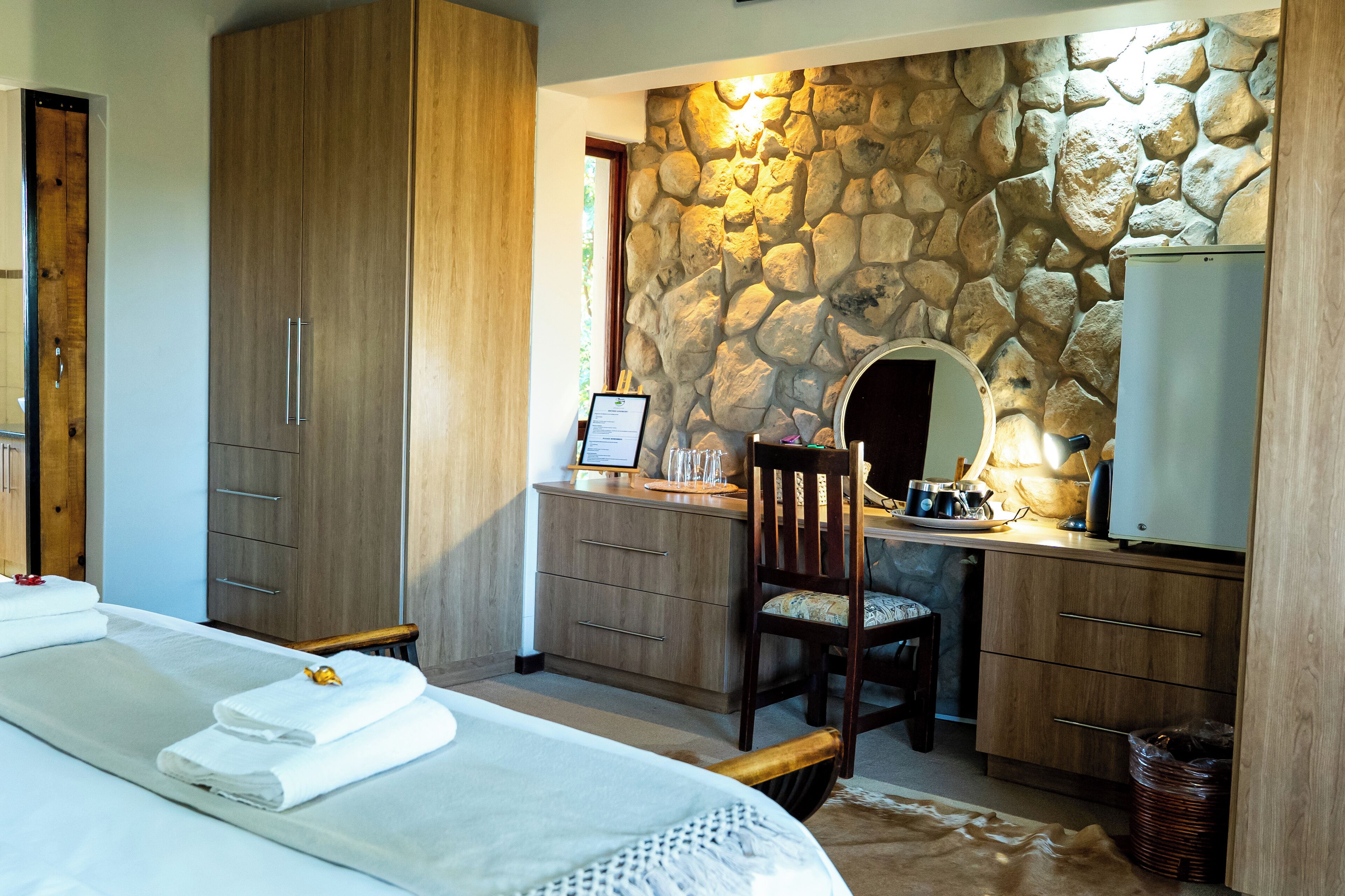 Une chambre avec un grand lit, une armoire en bois, un mur en pierre, un bureau avec une chaise et un miroir.