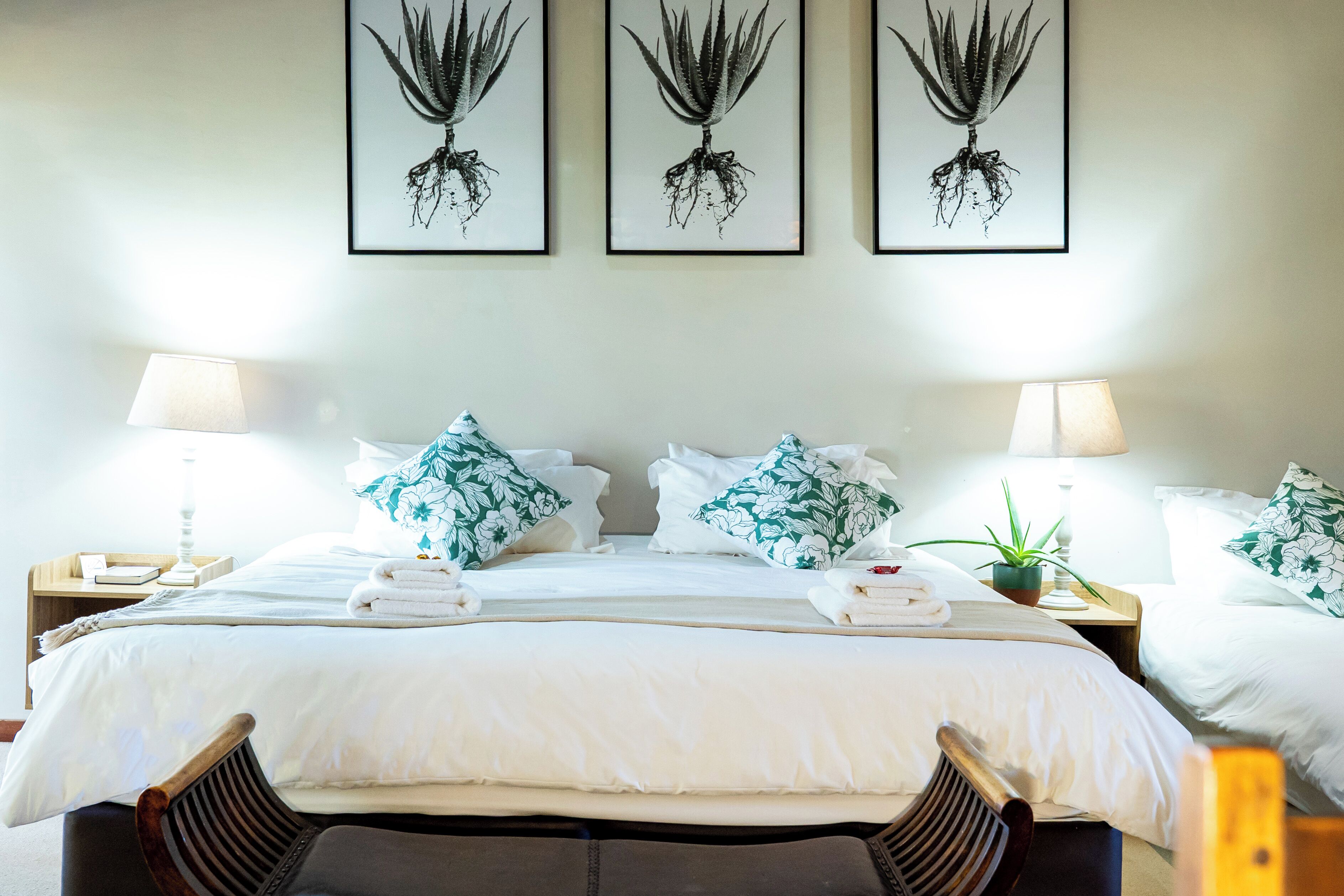 Una cama impecablemente arreglada con sábanas blancas y almohadas florales, flanqueada por mesitas de noche con lámparas y una pequeña planta en maceta.