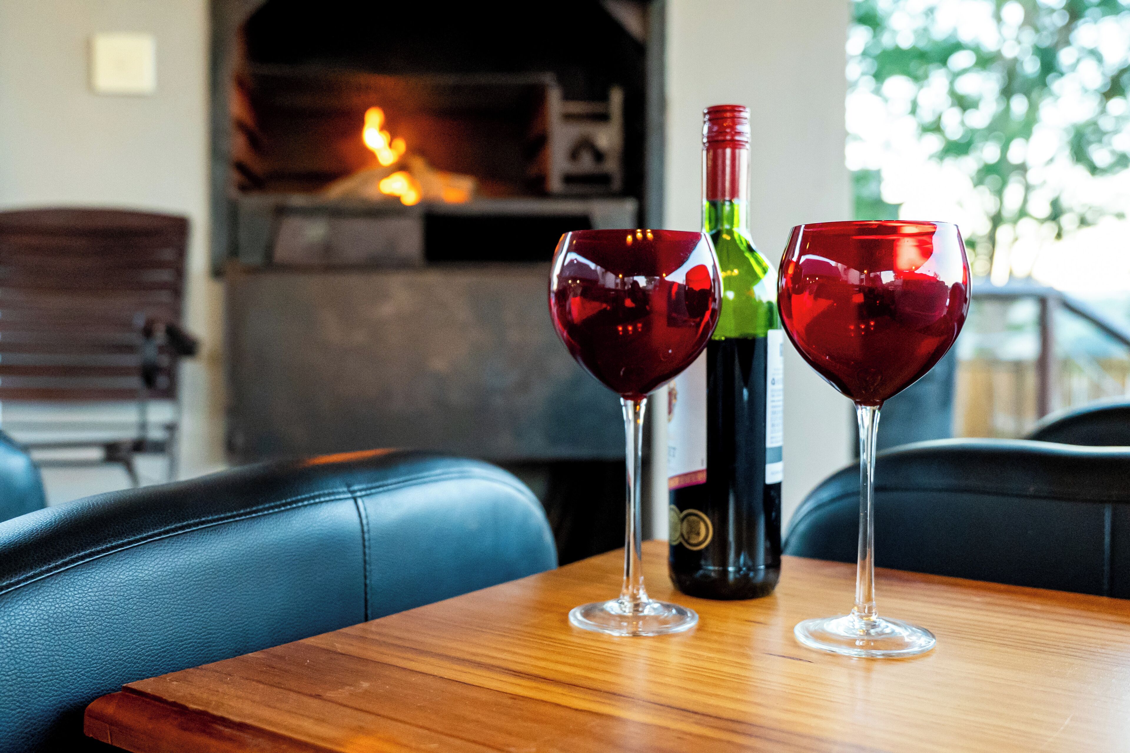 Two wine glasses on a wooden table with a bottle of wine, a fireplace, and a view of trees in the background.