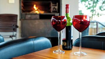 Two wine glasses on a wooden table with a bottle of wine, a fireplace, and a view of trees in the background.