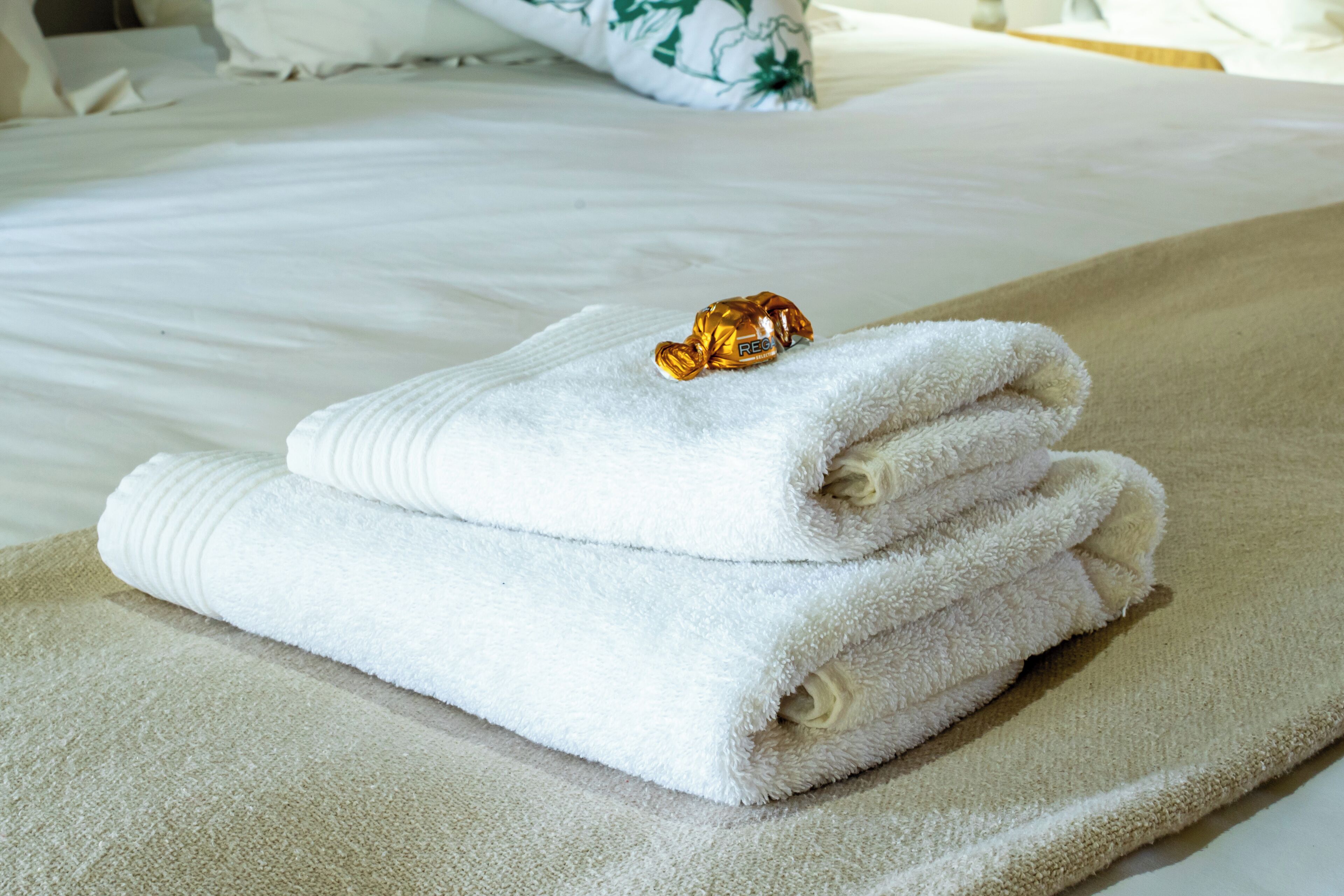A neatly folded white towel with a gold-colored object on top, placed on a beige towel on a bed with white bedding.