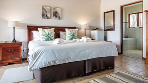 Uma cama bem arrumada com cabeceira de madeira, mesa de cabeceira com abajur e um banheiro visível por uma porta aberta.