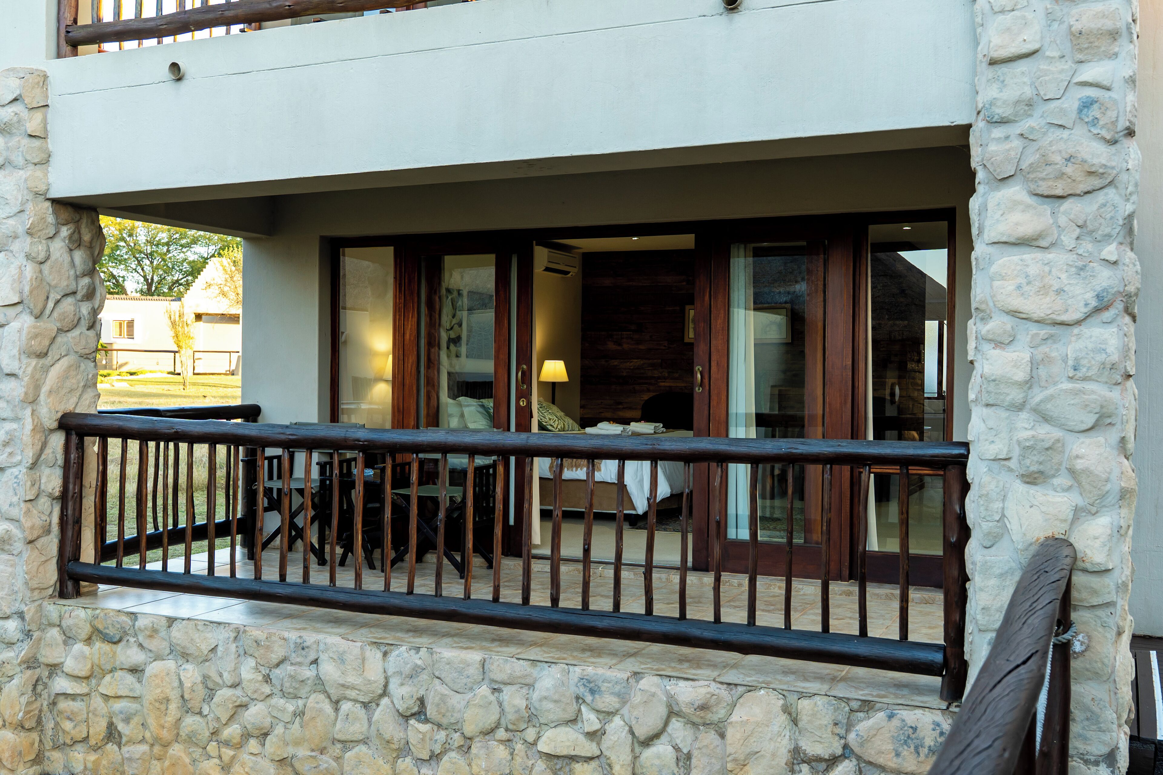 A stone-walled balcony with a wooden railing, a dining area with chairs and a table, and a view of a building with large windows.