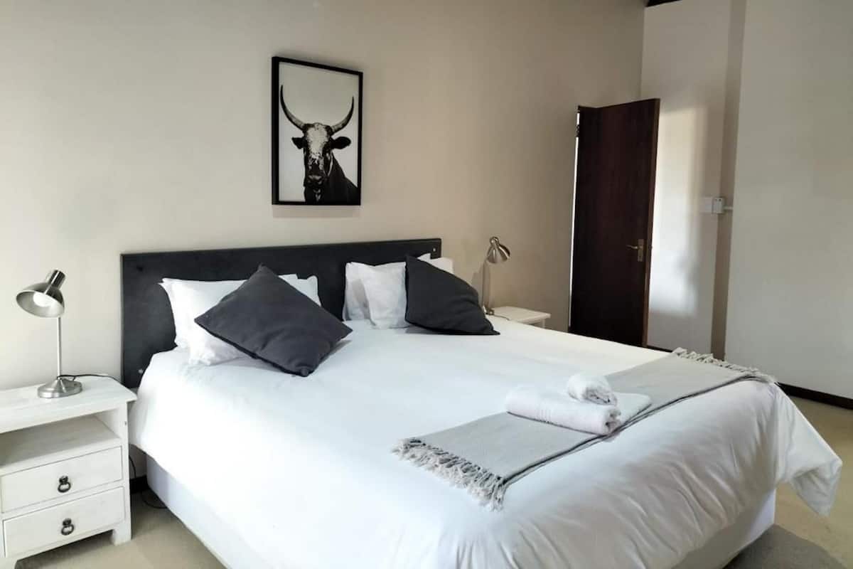 A neatly made bed with white linens and a gray throw blanket, a framed picture of a bull's head on the wall, and a wooden door in the background.