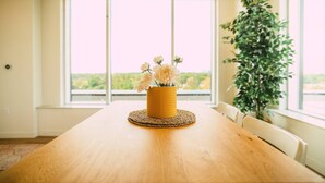 Een houten eettafel met een geweven placemat en een gele vaas met bloemen.
