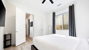 Un dormitorio con una cama grande, una ventana con cortinas, una puerta que da a otra habitación y una mesita.