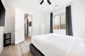Un dormitorio con una cama grande, una ventana con cortinas, una puerta que da a otra habitación y una mesita.