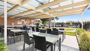 An outdoor patio area with tables and chairs, a brick wall, and a covered section with a metal roof. - Apollo Unit 1 - Ground Floor - A Block (Narooma)