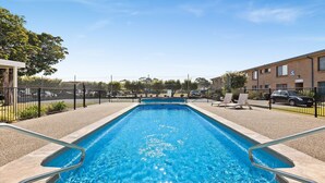 A rectangular swimming pool with a clear blue water, surrounded by a gravel area and a fence. There are lounge chairs and a building with a brick facade in the background. - Apollo Unit 1 - Ground Floor - A Block (Narooma)