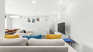 A modern living room with a sofa, a flat-screen TV, and a dining area with a wooden table and chairs. - Apollo Unit 1 - Ground Floor - A Block (Narooma)