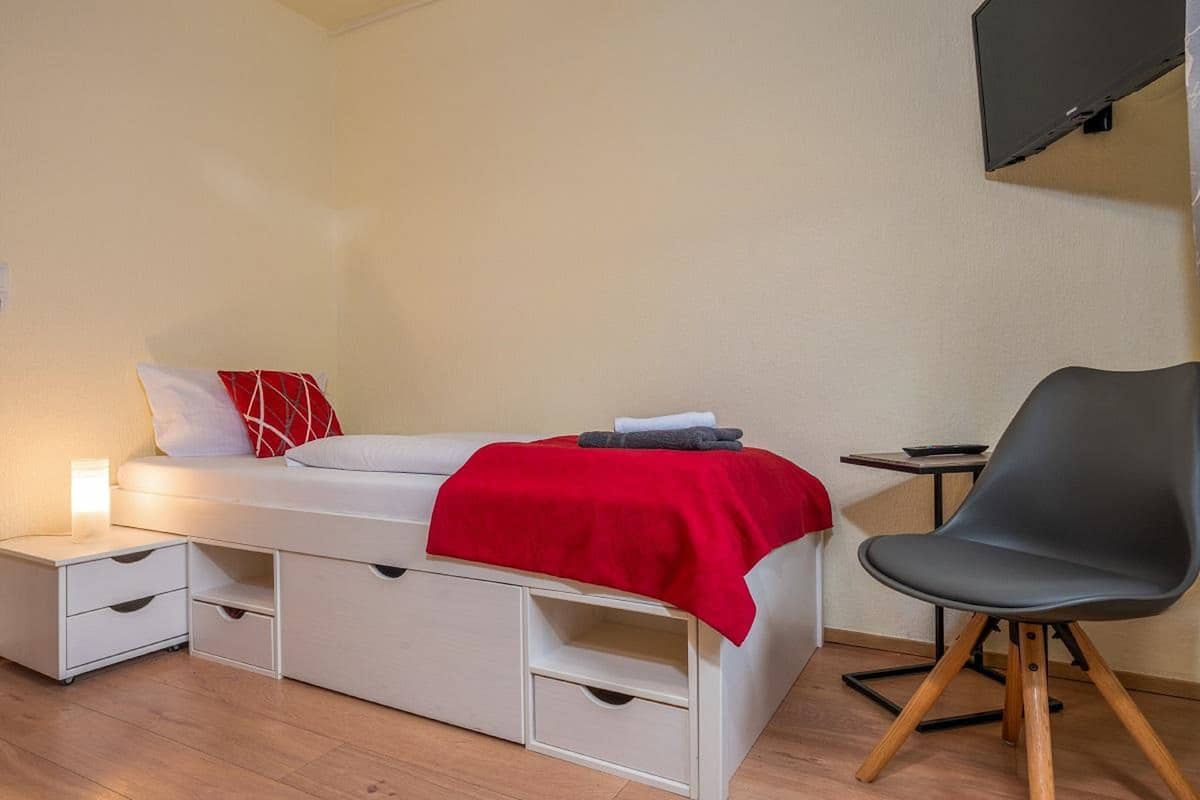 Une chambre à coucher de taille réduite, comprenant un lit, des tables de chevet, une chaise et un téléviseur fixé au mur.
