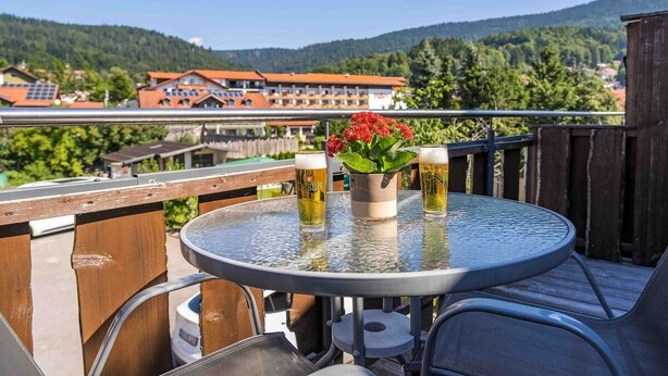 A round metal table with a flower vase and two beer glasses on a balcony overlooking a mountainous area with buildings in the background. - Pension Bayerwald (Bodenmais)