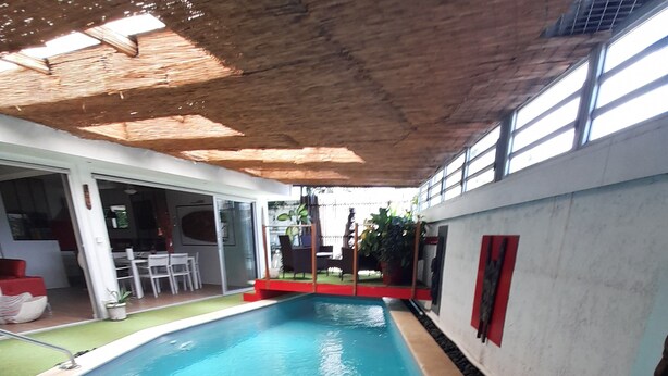 An indoor swimming pool with clear blue water, surrounded by a tiled deck and a covered area with a thatched roof. - Quartier Calme, Centre Ville, gde Chambre Piscine.................; (noumea)