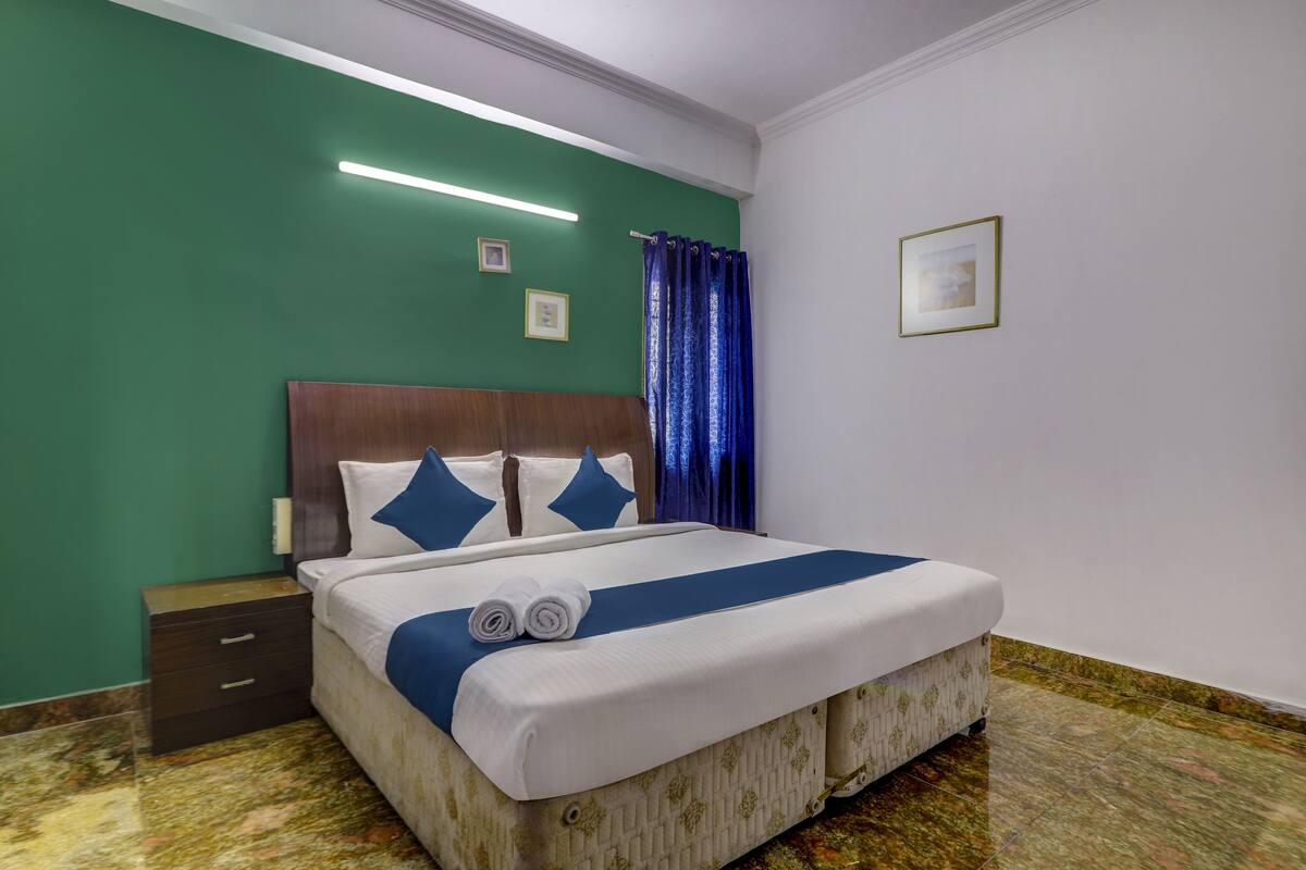 A neatly made bed with a blue bedspread and pillows, a wooden headboard, a nightstand with drawers, and a framed picture on the wall.