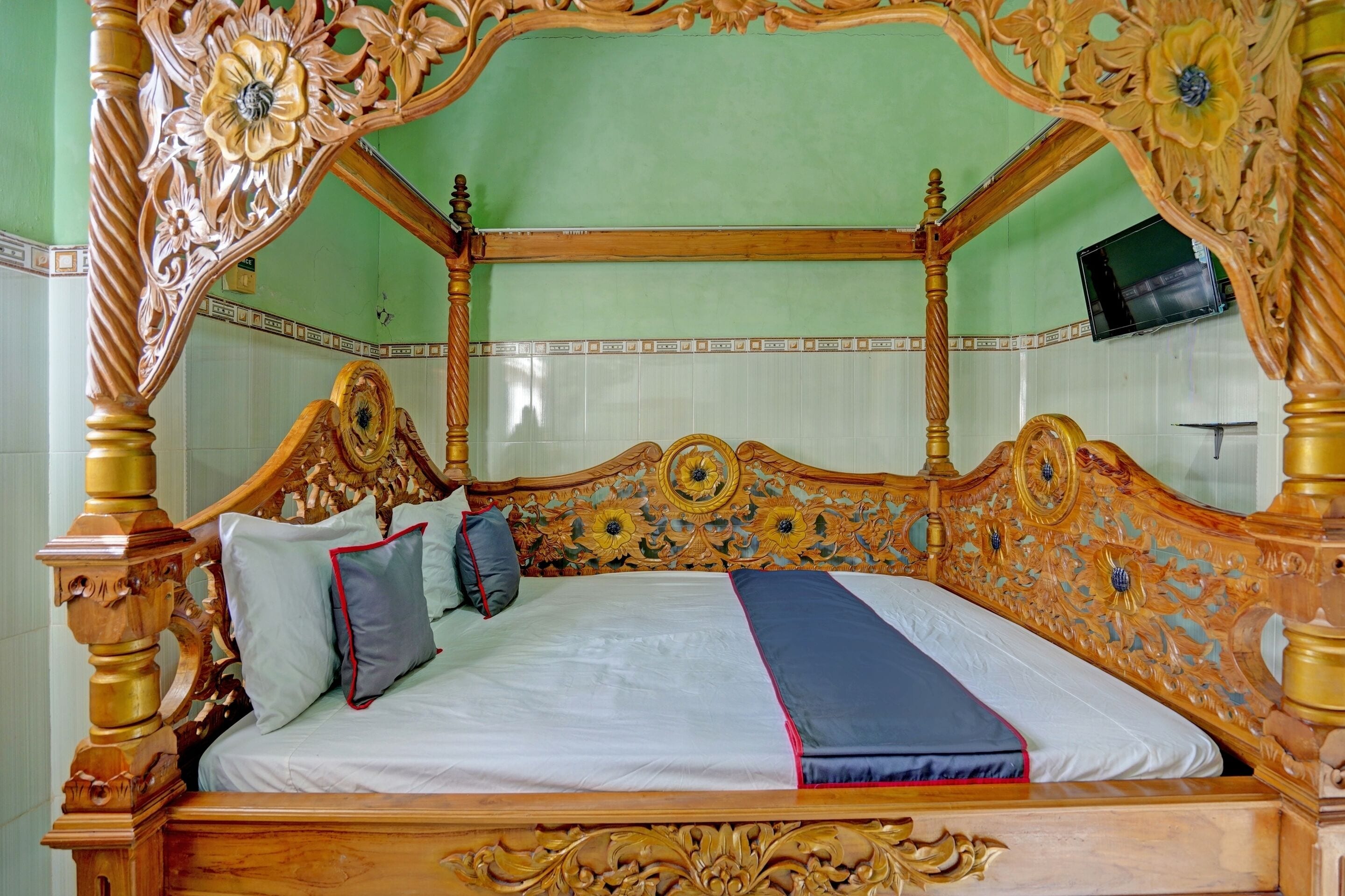A four-poster bed with intricate carvings, a television mounted on the wall, and a tiled floor.