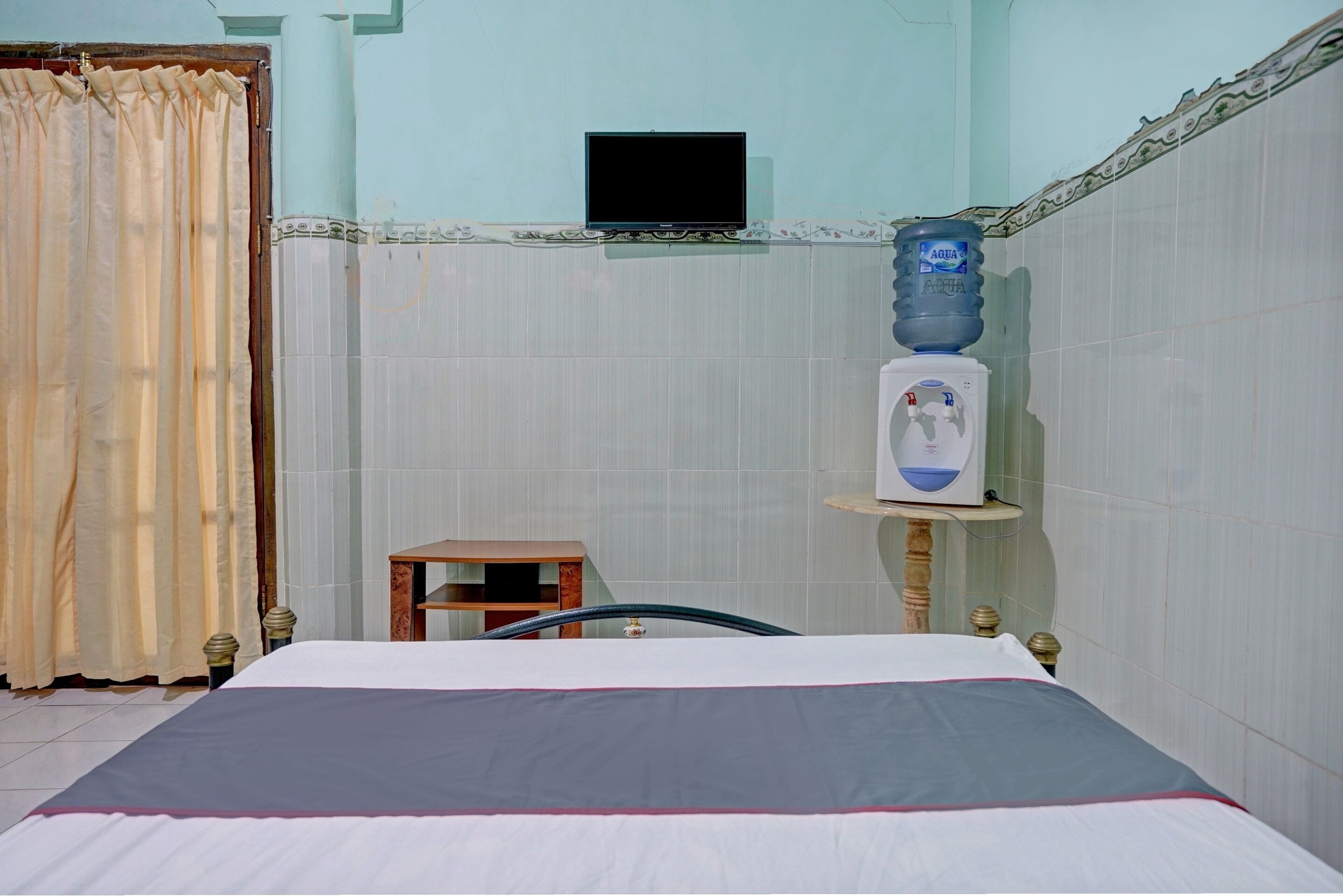 A hotel room with a bed, a water dispenser, a television mounted on the wall, and a curtain.