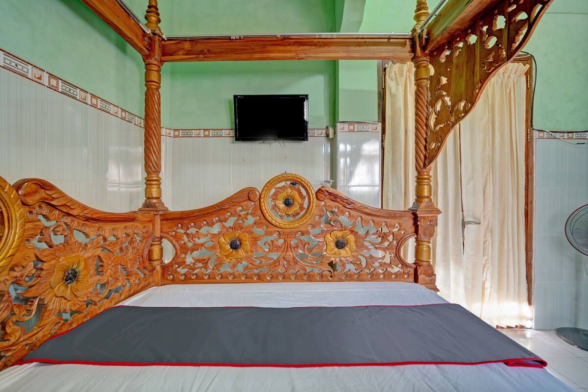 A four-poster bed with intricate floral carvings, a flat-screen TV mounted on the wall, and a fan on the floor.