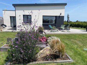 Een modern huis met een patio, straatmeubilair en een tuin vol planten en bloemen.