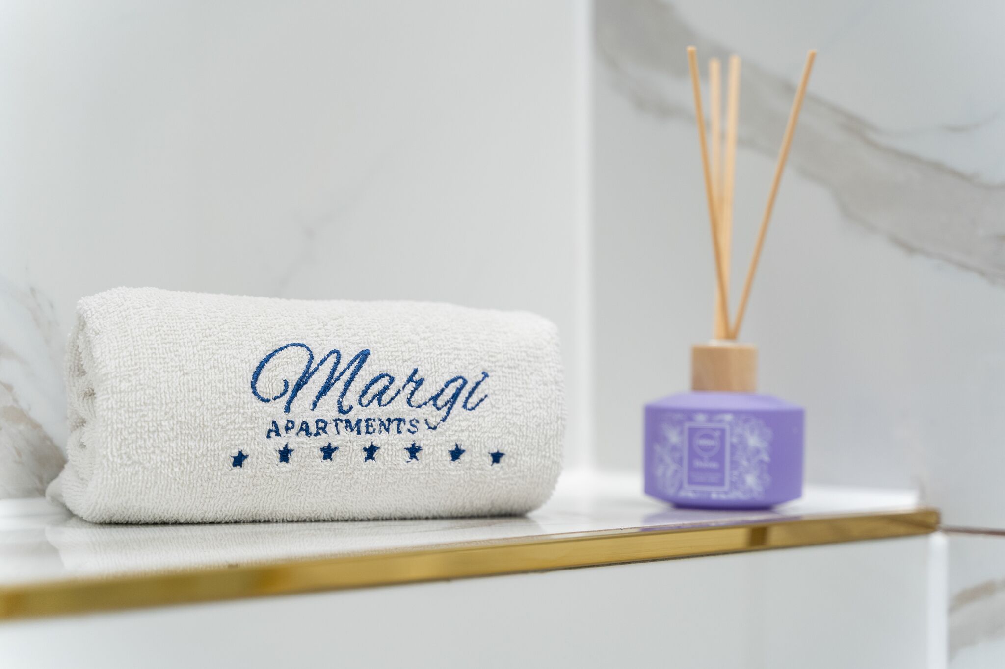 Valkoinen pyyhe, jossa on kirjailtu teksti ”Margi Apartments”, violetti huoneilmanraikastin ja taustalla marmoriseinä.