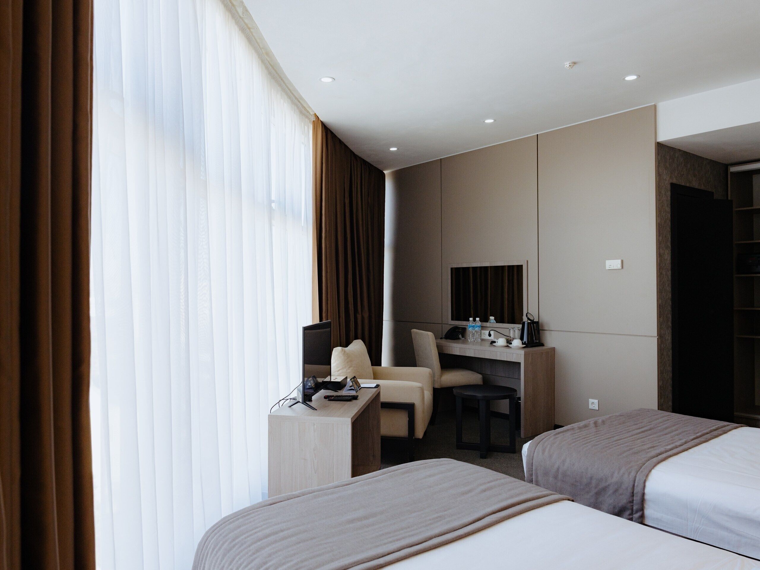 A hotel room with two beds, a desk with a chair, a small table, a mirror, and a window with curtains.