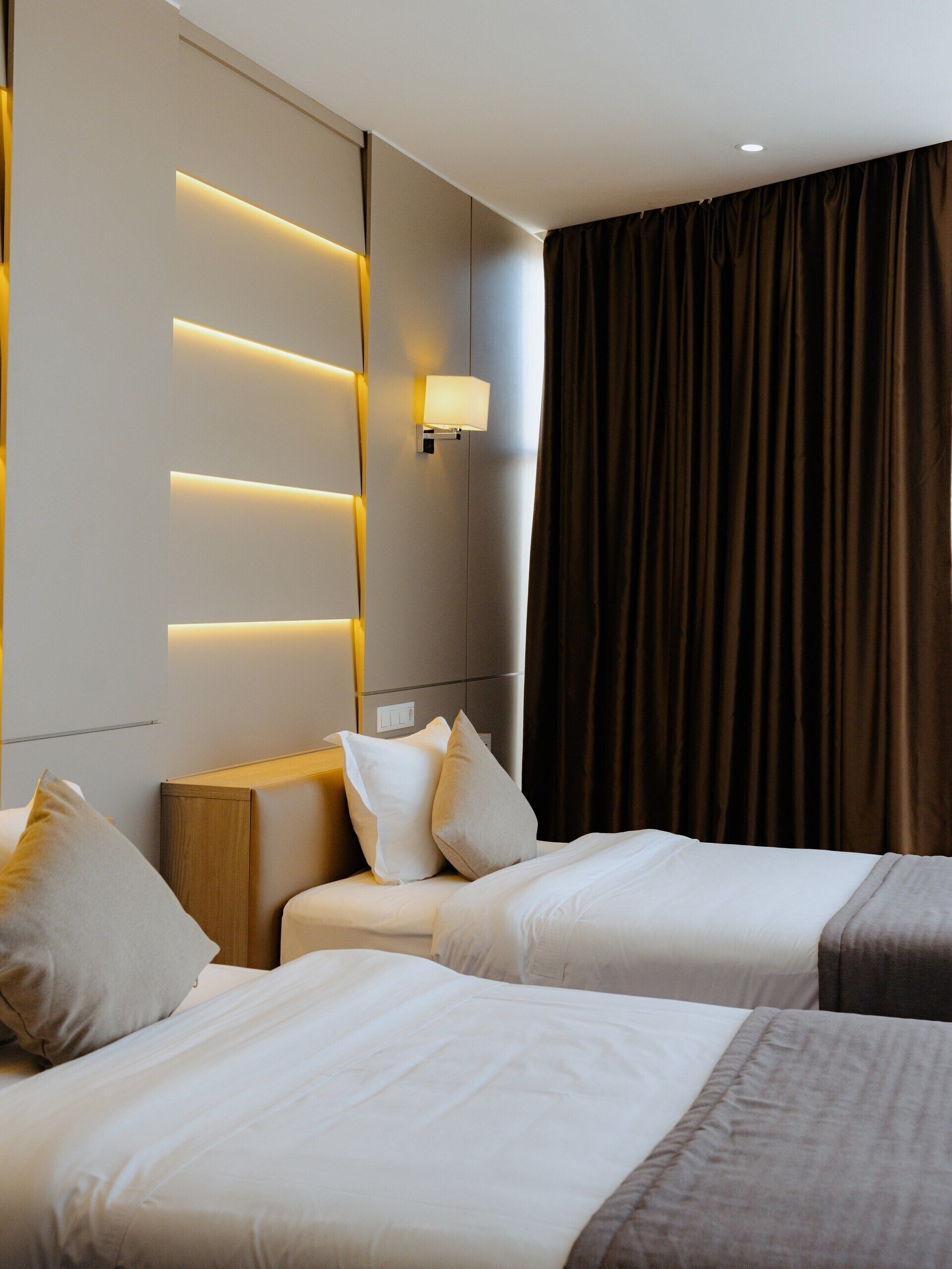 A hotel room with two beds, a bedside table, a wall-mounted light, and a large window with curtains.