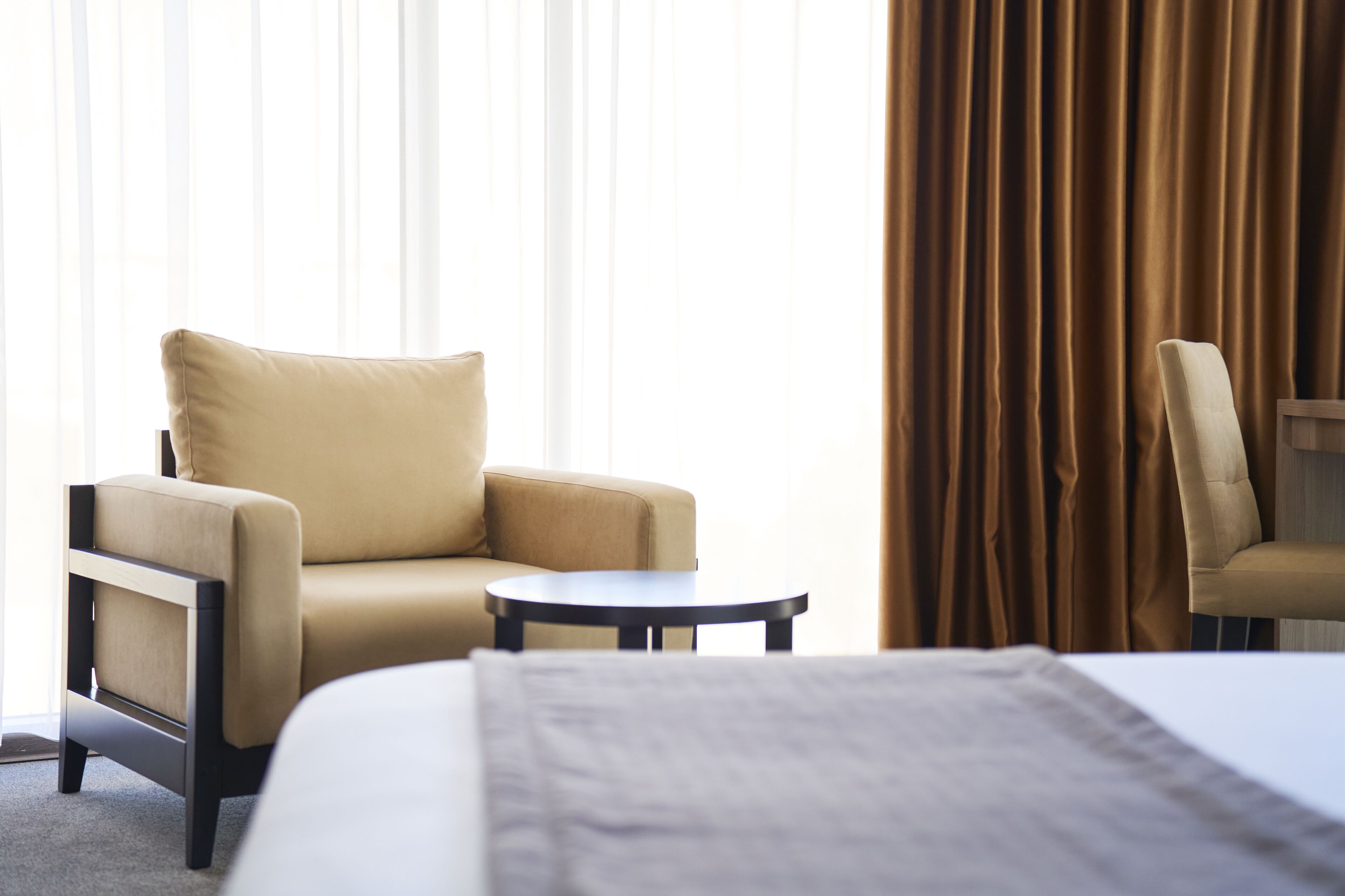 A hotel room with a bed, a beige armchair, a small round table, and a view of the outside through sheer curtains.