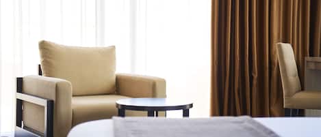 A hotel room with a bed, a beige armchair, a small round table, and a view of the outside through sheer curtains.