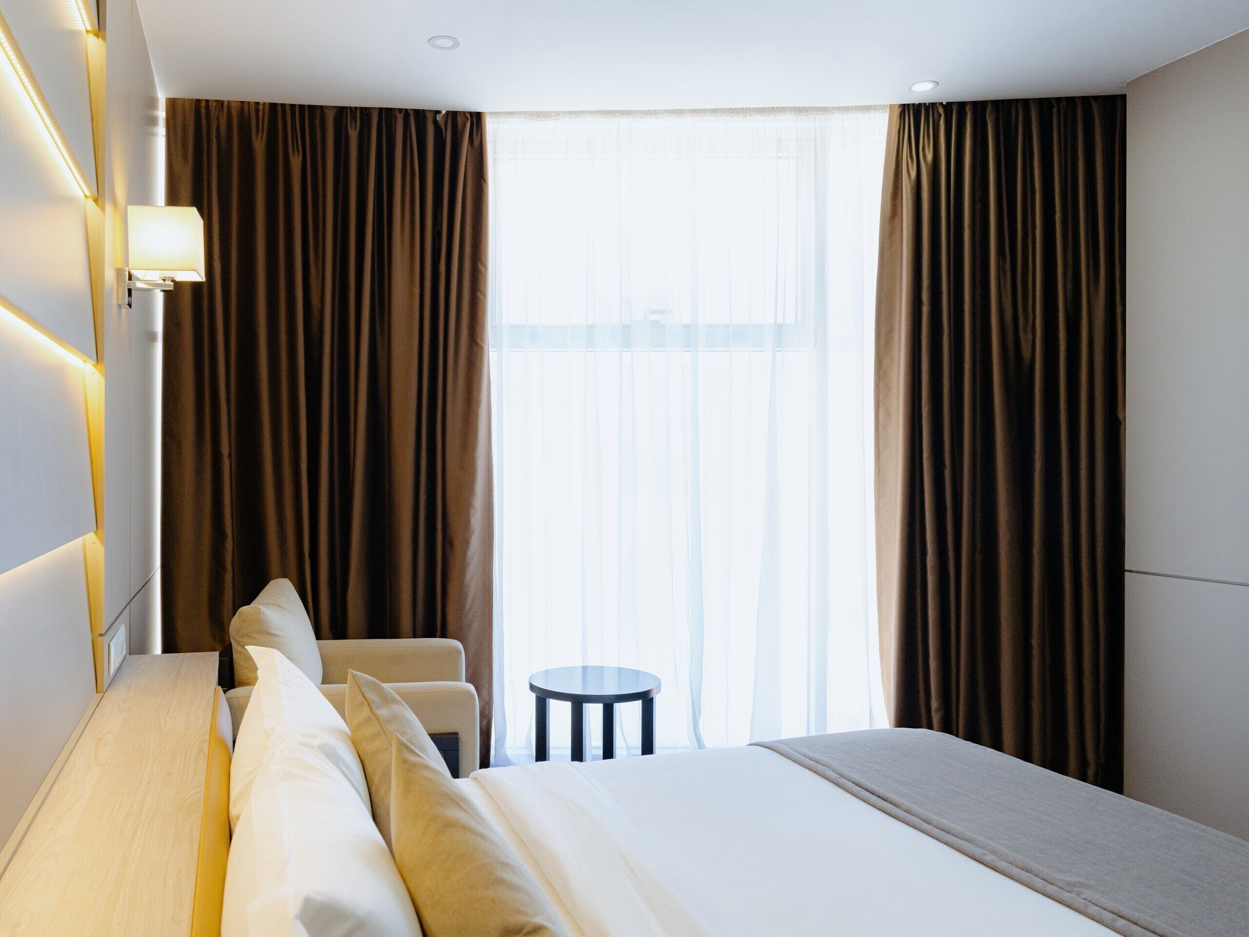 A hotel room with a bed, a bedside table, a chair, and a window with curtains.