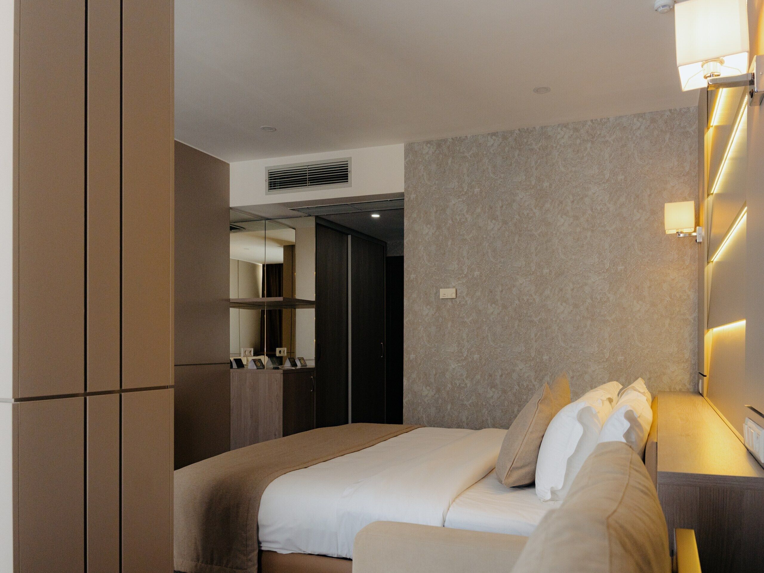 A modern hotel room with a large bed, a bedside table, a mirror, and a wall-mounted light fixture.
