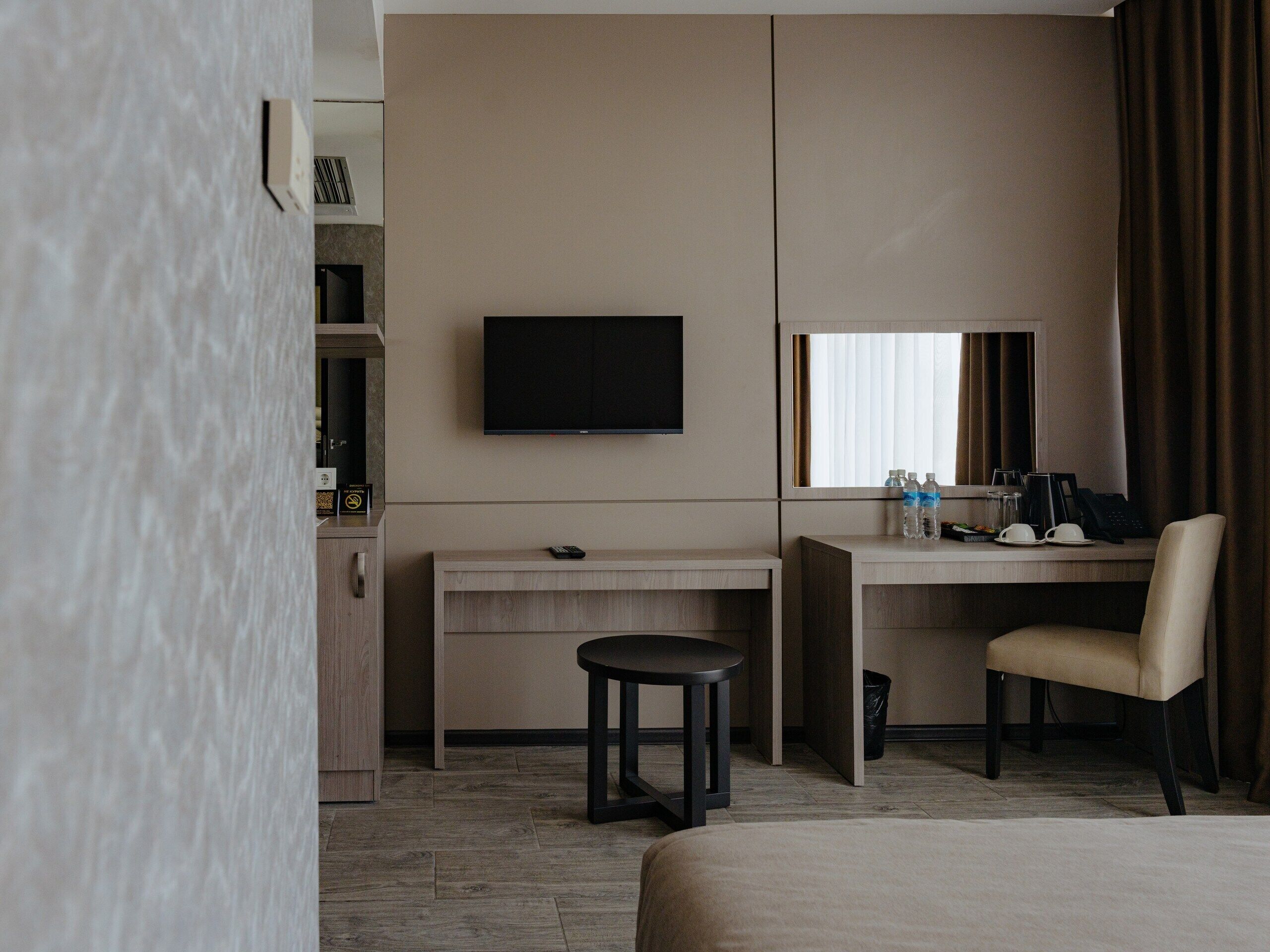 A hotel room with a bed, a desk with a chair, a television, a window with curtains, and a small stool.